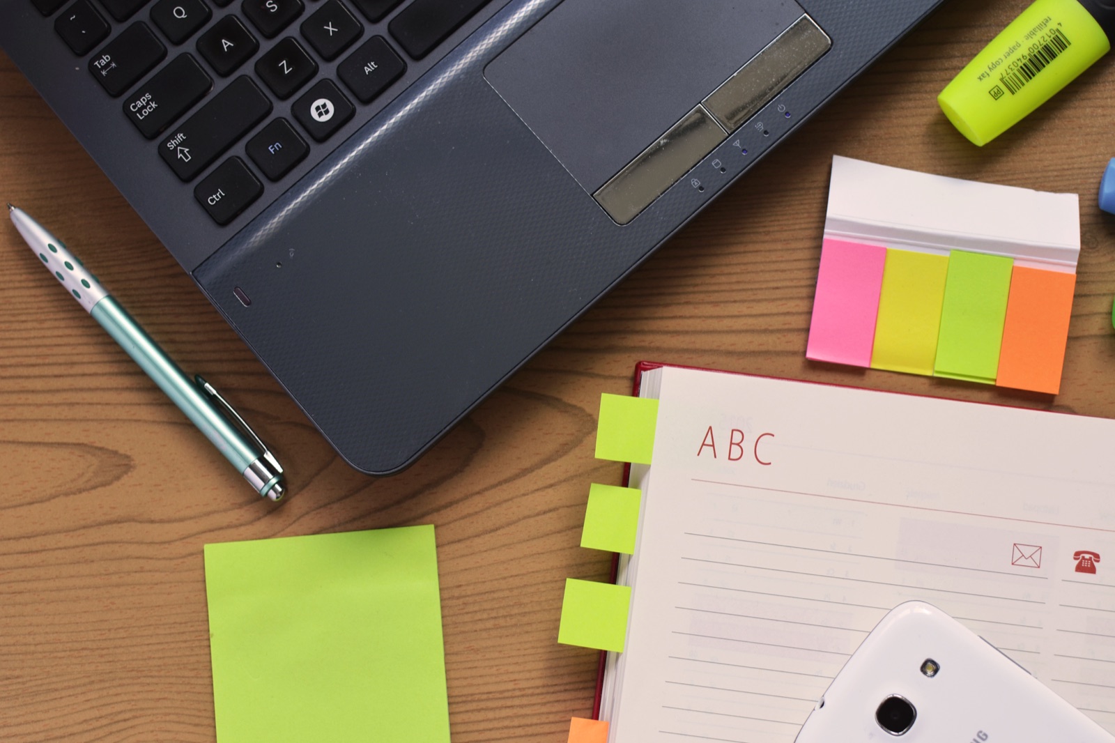 A laptop, pen, and open notebook on a wooden desk. Photograph by Pixel.la Free Stock Photos (CC0), via Wikimedia Commons.